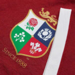 Close-up of red fabric featuring a shield crest with four quadrants (rose, shamrock, white emblem, red floral motif) and a white zipper edge.
