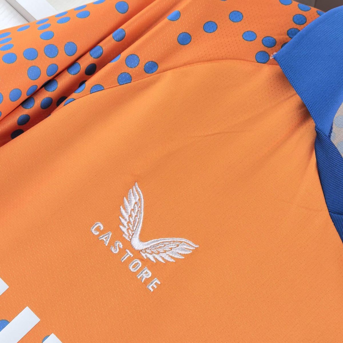 Close-up of an orange sports jersey with blue polka-dot accents and a white embroidered wing logo (Castore) showing partial text.