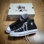 Black canvas high-top sneakers with white toe caps and laces, featuring a white star logo, placed on a wooden floor with a white branded box in the background.
