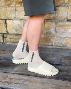 Person wearing beige ankle socks with vertical black text and white open-toe slide sandals, standing on a wooden deck in front of a stone wall.