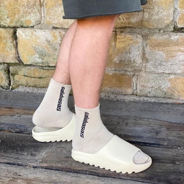 Person wearing beige ankle socks with vertical black text and white open-toe slide sandals, standing on a wooden deck in front of a stone wall.