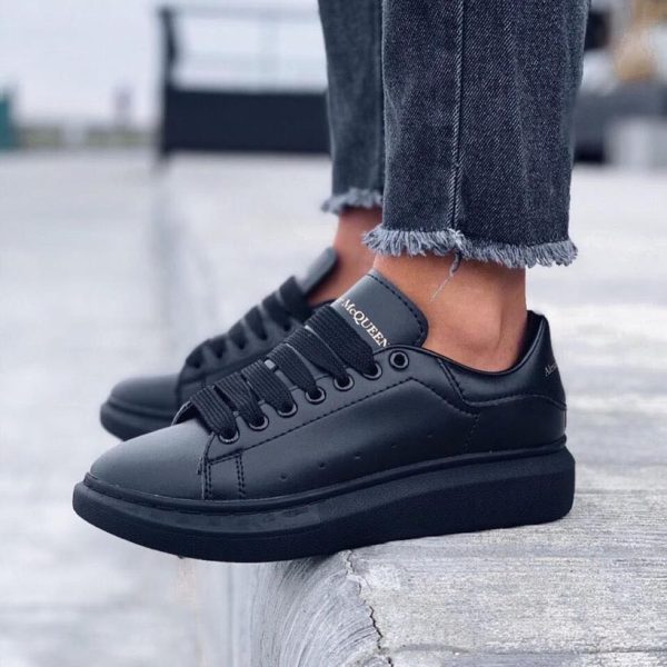Black leather low-top sneakers with thick sole and Jacquemus branding on the tongue, paired with frayed-hem jeans on a concrete ledge.