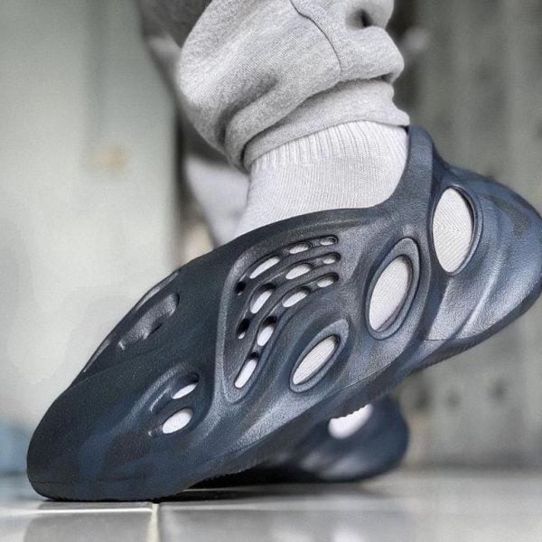 Close-up of a dark blue perforated slip-on shoe (Crocs-style) worn with a gray sock, stepping on a tiled floor.