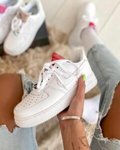 Person sitting with ripped jeans, holding a white Nike Air Force 1 sneaker; neon green nail visible on the thumb; other white sneakers in the background.