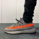 Grey and orange Adidas Yeezy Boost 350 V2 sneaker with an orange stripe and 'SPLY-350' text, worn with black pants on a carpeted floor