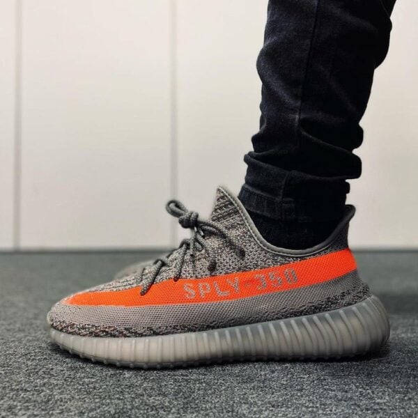 Grey and orange Adidas Yeezy Boost 350 V2 sneaker with an orange stripe and 'SPLY-350' text, worn with black pants on a carpeted floor
