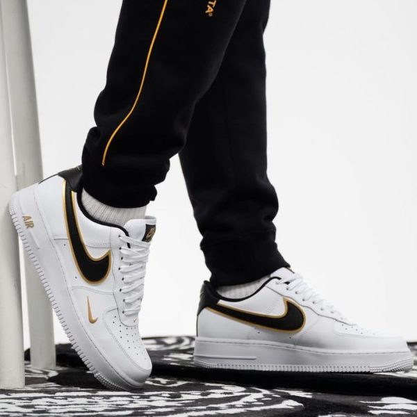 Person wearing white Nike Air sneakers with black and gold swoosh, standing on a patterned rug; Nocta branding on black pants with orange stripe.