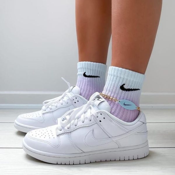 Person wearing white Nike sneakers with pastel gradient socks and a gold ankle chain with a blue heart charm.