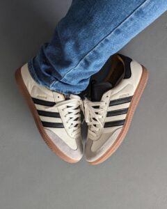 White Adidas-style sneakers with black stripes and gum soles, paired with blue jeans on a gray floor (top-down view).