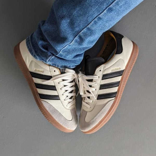 White Adidas-style sneakers with black stripes and gum soles, paired with blue jeans on a gray floor (top-down view).