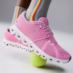 Pink athletic running shoe stepping on a bright tennis ball, with striped rainbow socks visible behind it.