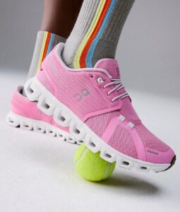27a68a48-51d6-498c-a535-b993a1f614d0 | UAE SPORT STORE Pink athletic running shoe stepping on a bright tennis ball, with striped rainbow socks visible behind it.