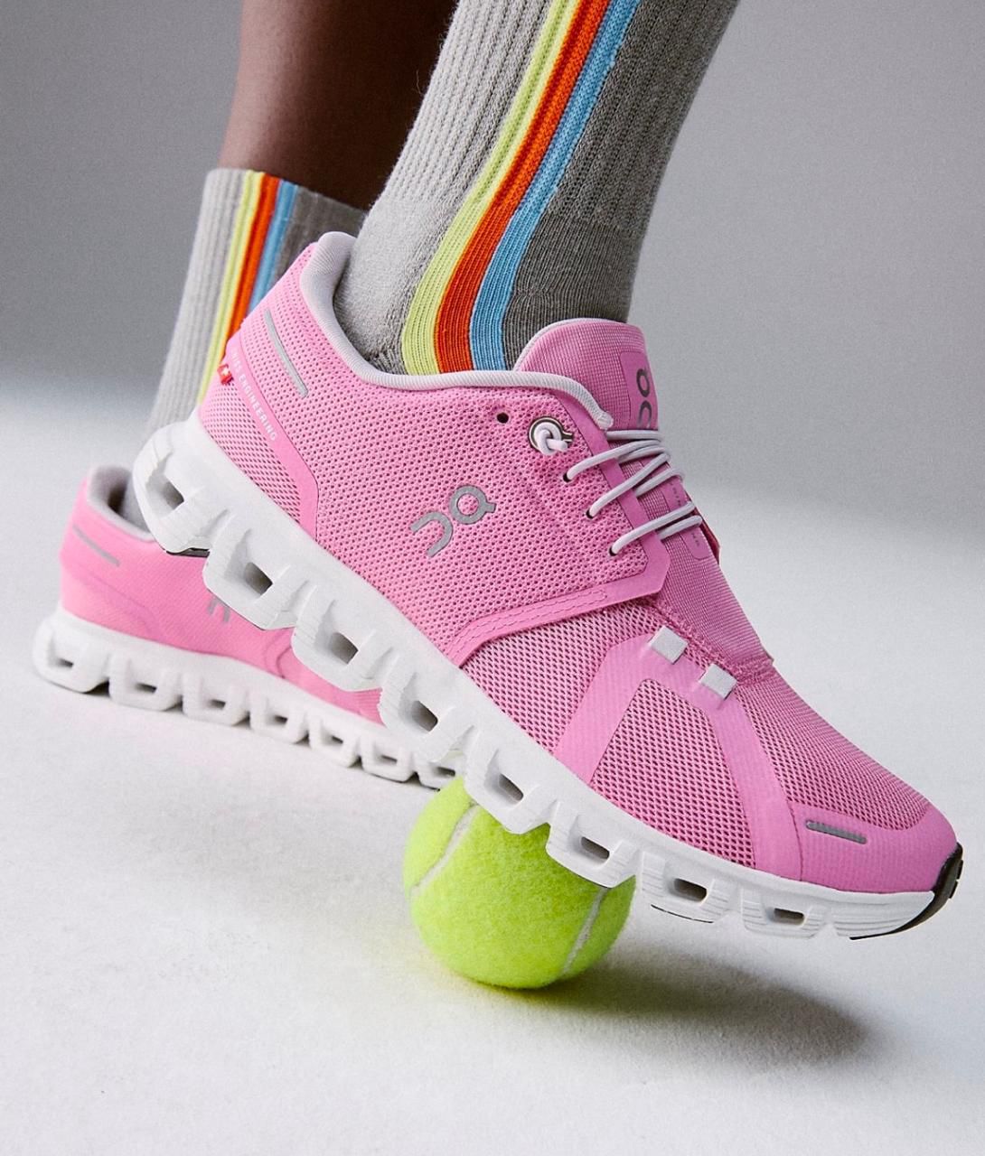 27a68a48-51d6-498c-a535-b993a1f614d0 | UAE SPORT STORE Pink athletic running shoe stepping on a bright tennis ball, with striped rainbow socks visible behind it.