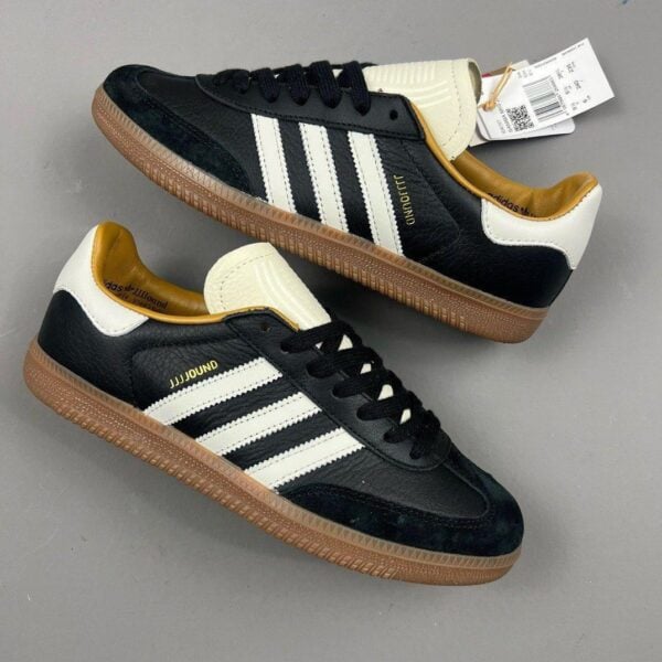 Black leather sneakers with white Adidas stripes and light brown gum soles, showing a tag from a0 collaboration.