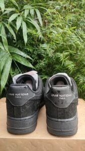 Two black Louis Vuitton sneakers with LV logo on the back, placed on a cardboard box outdoors with green plants behind.