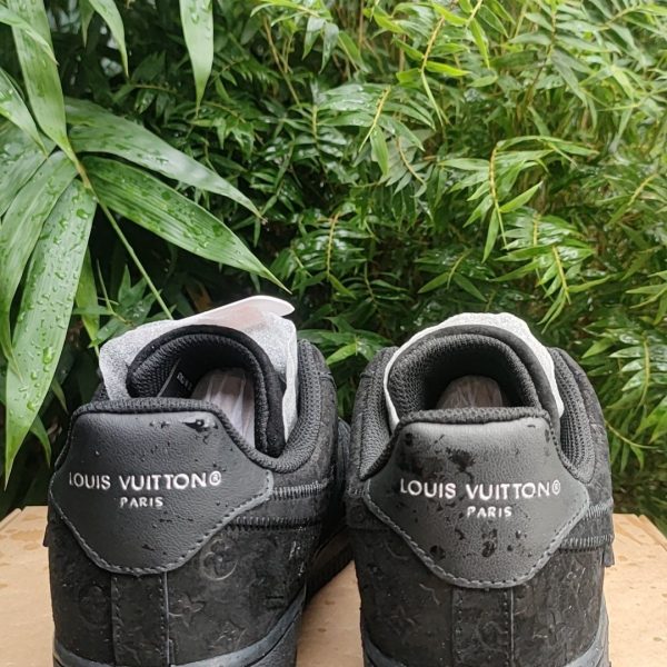 Two black Louis Vuitton sneakers with LV logo on the back, placed on a cardboard box outdoors with green plants behind.