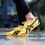 Close-up of yellow sneakers with dark stripes as the wearer takes a step on a glossy tiled surface, mid-motion.