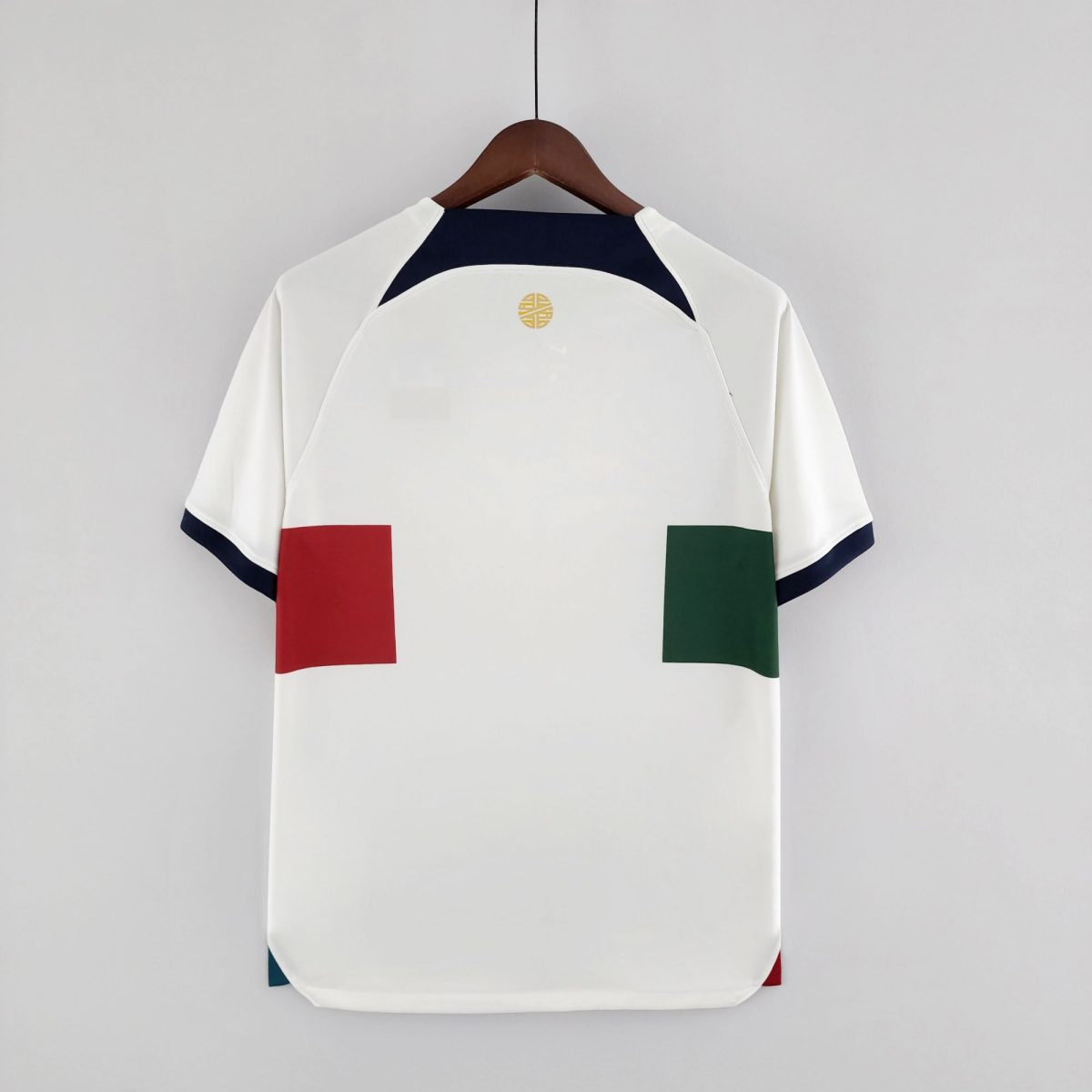 Back view of a white short-sleeve jacket on a wooden hanger, featuring a dark navy yoke, small gold circular logo, and red and green color blocks at the lower sides against a gray wall.