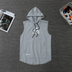 Flat lay of a gray sleeveless hooded top with drawstrings and a front kangaroo pocket on a dark wooden surface. Includes Under Armour logo near the pocket edge.
