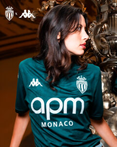 Woman in a teal AS Monaco football jersey looks to the right, standing beside a metallic sculpture.