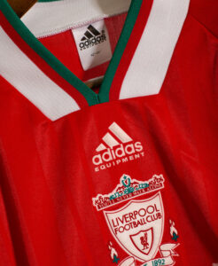 Close-up of a red Liverpool FC jersey showing the white Adidas logo and crest, with a green-and-white collar, hanging on a hanger.