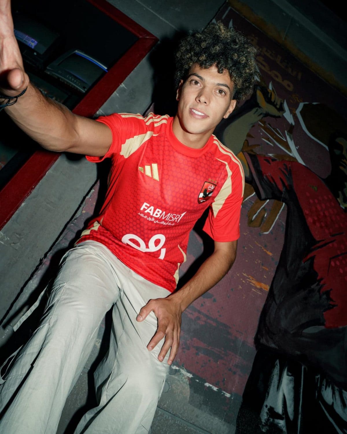 Young man with curly hair in a red athletic jersey taking a selfie in a graffiti-covered hallway, arm extended toward the camera.