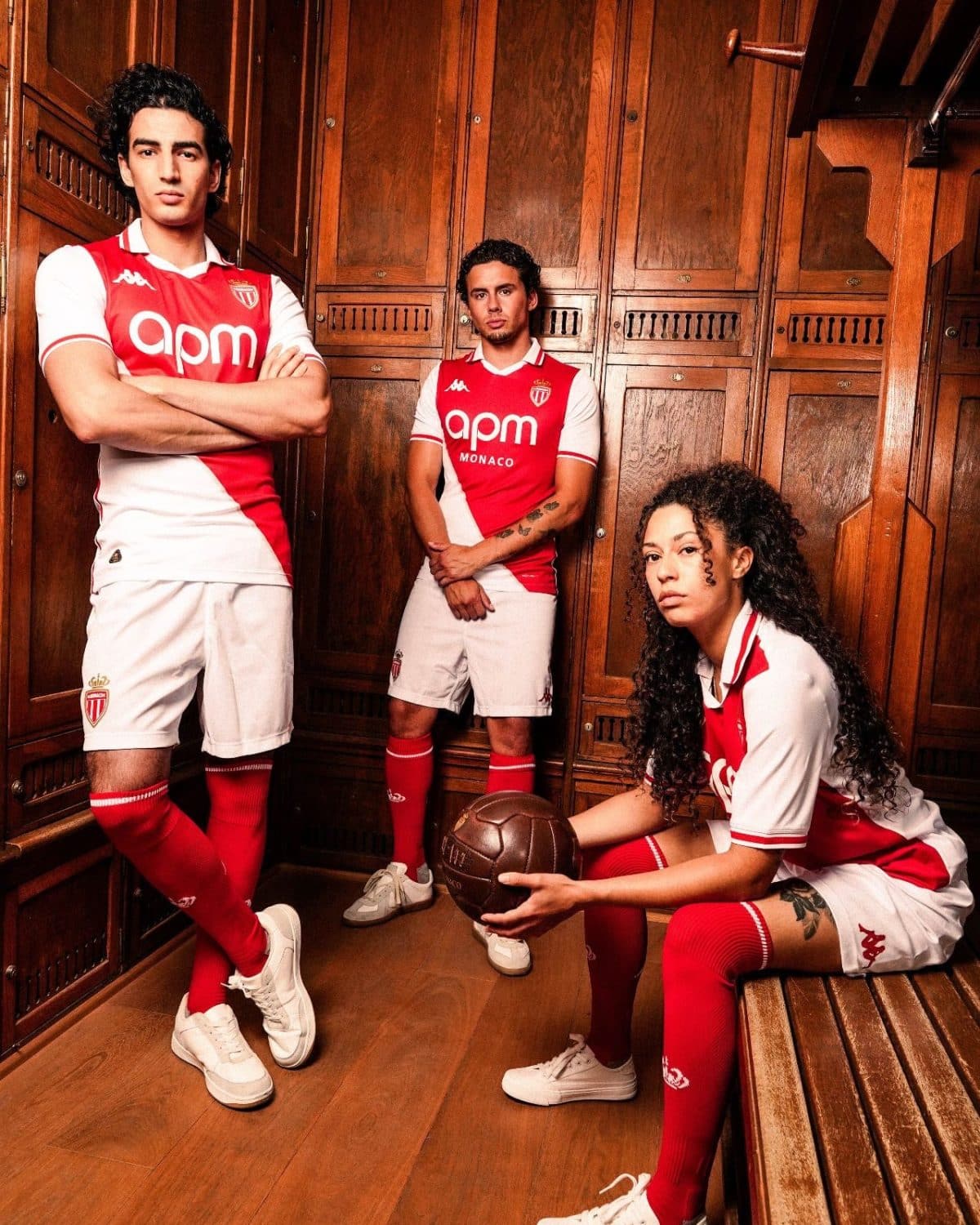 Three soccer players in red-and-white Monaco kits pose in a wooden locker room, one standing with arms crossed, one standing with hands clasped, and a seated player holding a ball.