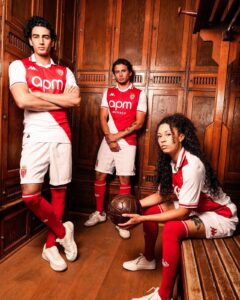 2Rpt82dAGwTuLj4 | UAE SPORT STORE Three soccer players in red-and-white Monaco kits pose in a wooden locker room, one standing with arms crossed, one standing with hands clasped, and a seated player holding a ball.