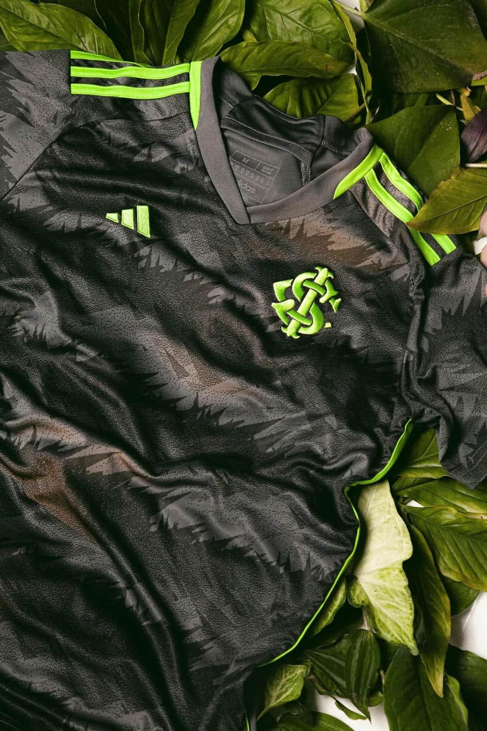 2SzdHbMmUBn0ySH_b0588015-7b32-4dfb-aa7b-c0e48b9a30e0 | UAE SPORT STORE Close-up of a black football jersey with bright neon green accents, Adidas logo, and a neon green club crest, laid on green leaves.