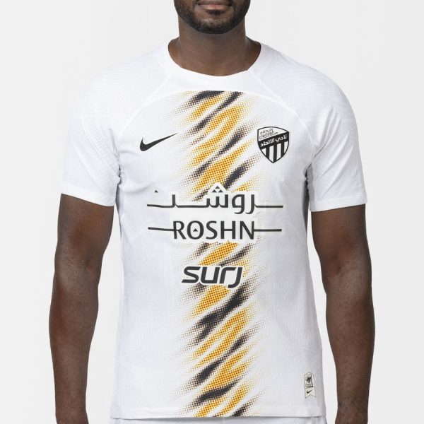 Male model wearing a white soccer jersey with a diagonal orange-black pattern, ROSHN branding, and a team crest on the chest.