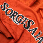 Orange fabric with bold black-and-white outlined letters spelling 'SORGESANA' laid diagonally across the material, showing a textured sportswear look.