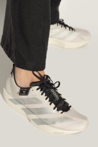 Close-up of a light-gray Adidas sneaker with black laces worn with black pants, showing the side stripes and textured upper.