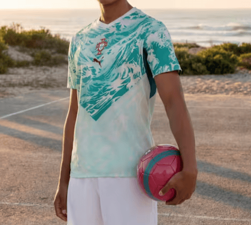 2_40700202-ac61-4093-b825-78ef0db3965e | UAE SPORT STORE Young man in a teal patterned soccer jersey holds a pink and blue soccer ball at a sandy beach during sunset.