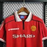 Red Manchester United football jersey on a wooden hanger, featuring the Umbro logo, club crest, and SHARP sponsor text.