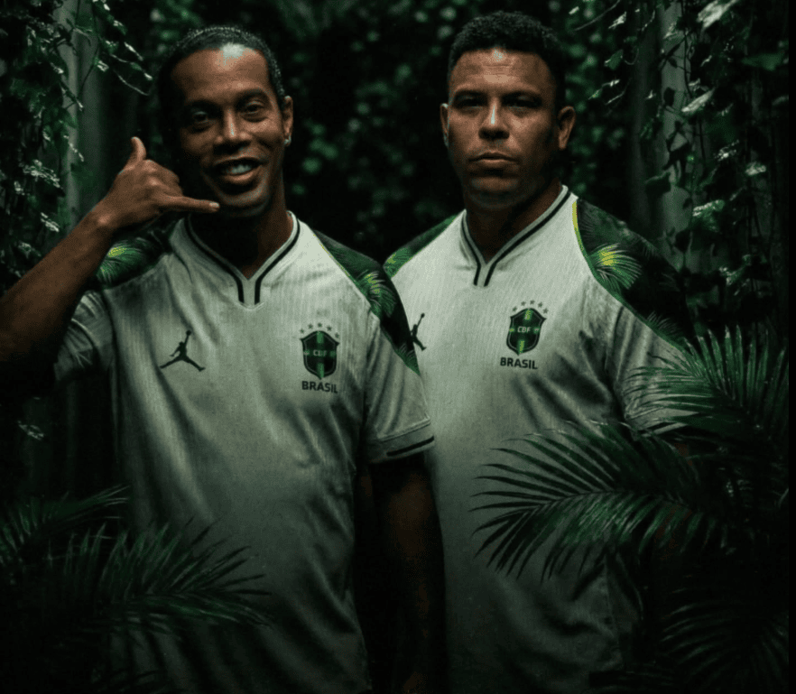 2_a908043f-9046-4f8f-a93f-96b5f90aa189 | UAE SPORT STORE Two men in Brazil soccer jerseys stand among dense jungle foliage, looking at the camera, one flashing a shaka sign with his hand.