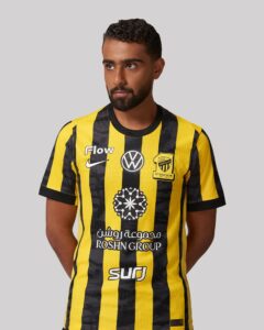 Man wearing a yellow and black striped soccer jersey with sponsor logos, facing slightly to the right against a gray background.