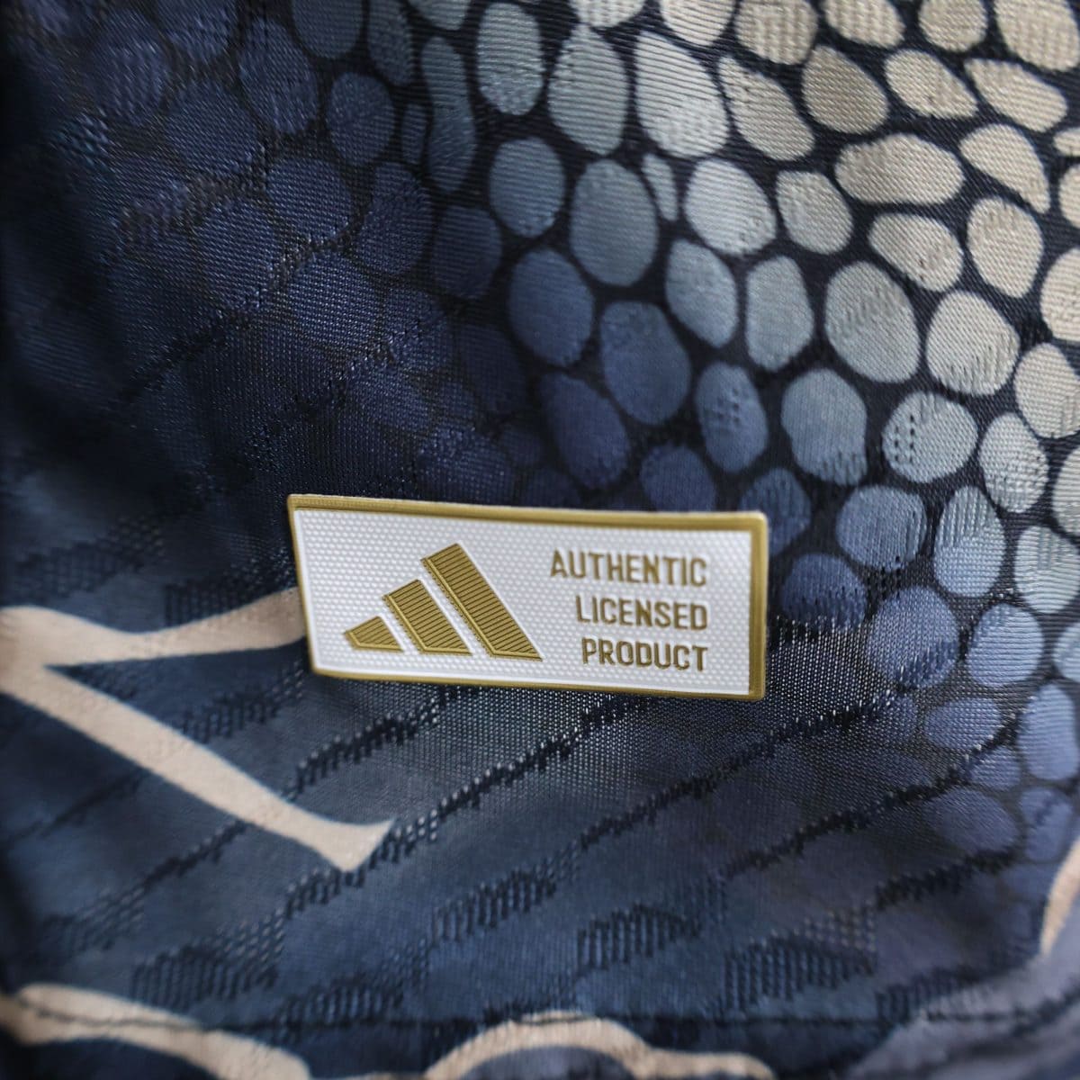 Close-up of a badge on dark patterned fabric, showing text 'AUTHENTIC LICENSED PRODUCT' and gold diagonal stripes.
