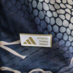 Close-up of a badge on dark patterned fabric, showing text 'AUTHENTIC LICENSED PRODUCT' and gold diagonal stripes.