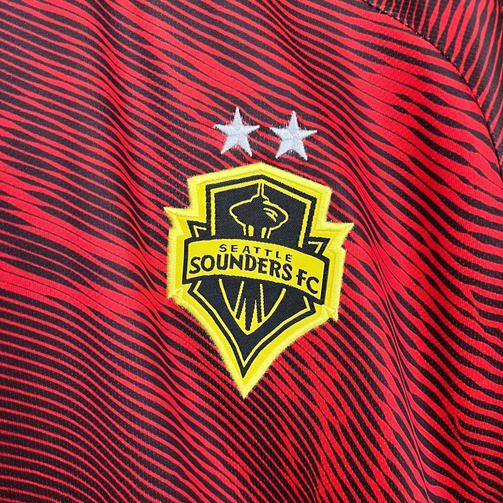 2b4044a5_3c46375d-2b80-4fb8-a437-45c3a16e71db | UAE SPORT STORE Sewn Seattle Sounders FC shield patch on red-and-black striped fabric with two white stars above the patch area.