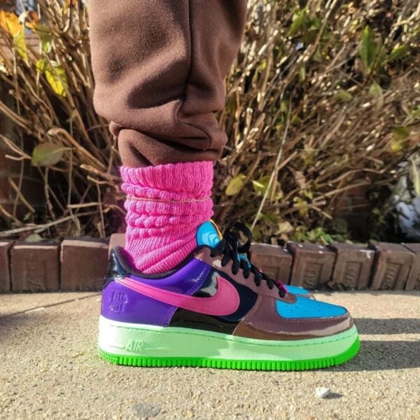 Close-up of a colorful Nike sneaker with neon green sole, pink swoosh, and blue toe, worn with a pink knit sock on a sidewalk.