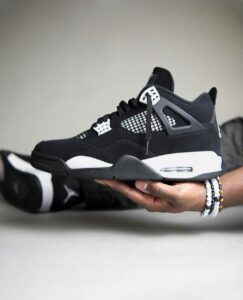 Side view of a black and white sneaker (Air Jordan 4) being held horizontally by a hand wearing white beaded bracelets on the wrist.