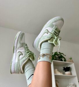 Mint green suede sneakers with white soles and laces, worn with light blue ribbed socks, indoors near a plant-filled shelf.