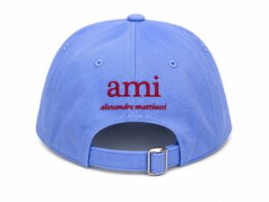 Back view of a light blue baseball cap with a metal buckle strap and red embroidered text 'ami' followed by 'alexandre mattiussi' on the back panel.