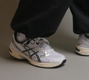 2ce7d161-1fe8-48b2-aaae-83b7e87982de | UAE SPORT STORE Pair of white athletic sneakers with black stripes on a gray floor, worn with black pants.