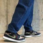 Casual sneakers with a green Nike swoosh, camo pattern panels, and white midsoles, worn with blue jeans beside a concrete wall.