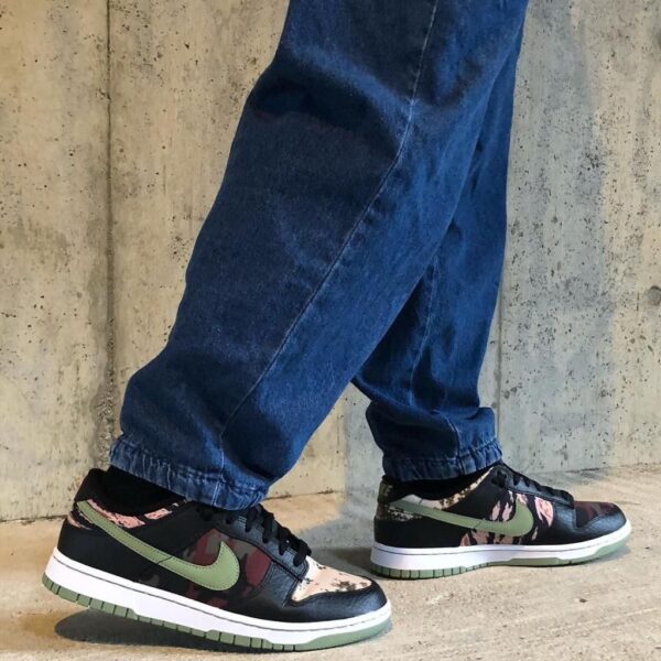 Casual sneakers with a green Nike swoosh, camo pattern panels, and white midsoles, worn with blue jeans beside a concrete wall.