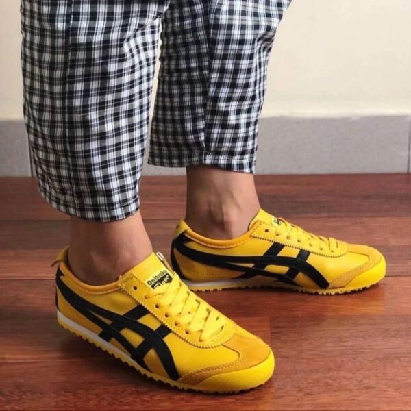 Person wearing bright yellow Onitsuka Tiger sneakers with black side stripes, standing on a wood floor in plaid pants.