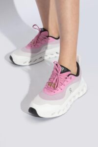 2d7ce056-279d-4a81-9881-8202c1d472fe | UAE SPORT STORE Pink and white athletic sneakers on a person’s feet with a chunky white sole and pink laces against a light gray background, styled for sport or fashion.