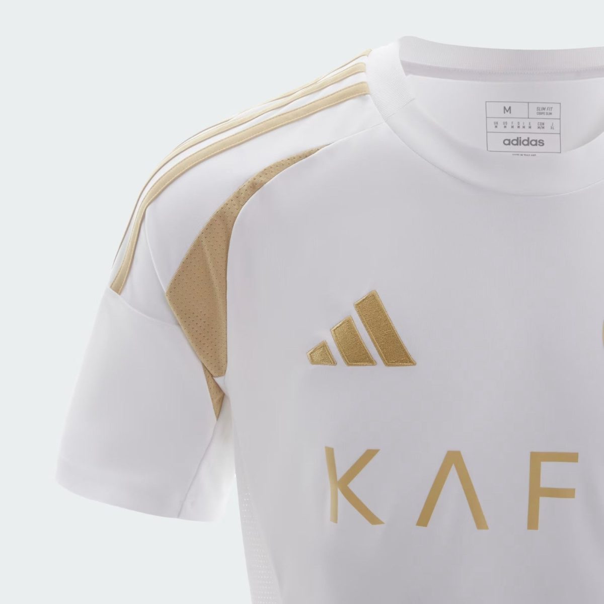 White Adidas sports jersey close-up showing gold shoulder stripes and a gold emblem, with partial gold letters visible (KAE).