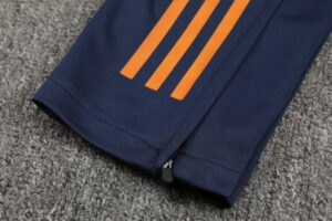 Close-up of navy athletic pants leg with three orange stripes and a cuff zipper on a gray carpet surface